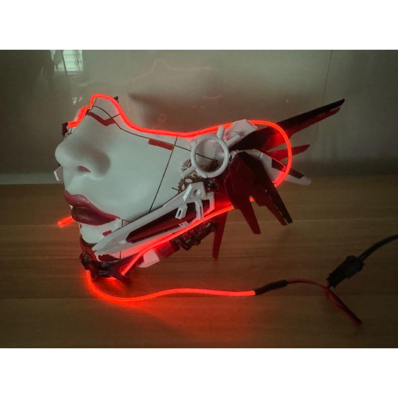 Może przedstawiać: Biała i czerwona plastikowa maska z czerwonym świecącym światłem. Maska ma ludzką twarz z detalicznym wzorem.