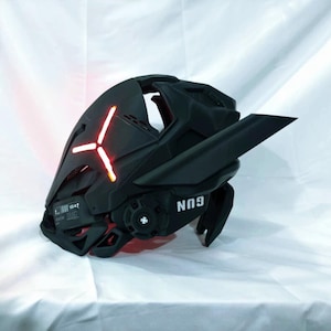 Op de afbeelding: Zwarte futuristische helm met een rood verlicht kruisvormig ontwerp aan de voorkant. De helm heeft hoekige kenmerken, een vizier en het woord "GUN" aan de zijkant gedrukt. De helm is waarschijnlijk voor cosplay of een thema-evenement.