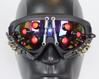 May include: Black ski goggles with red LED lights and silver accents. The goggles have a spiked frame and are worn on a black mannequin head.