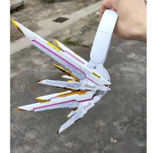 May include: A white and gold model with pink accents, resembling wings or a futuristic design. The model has sharp, angular elements and text that reads "ST75-802". The design is held by a hand, showcasing its size and detail.