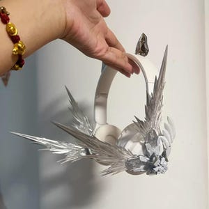 Può includere: Cuffie bianche con decorazioni argentate a forma di ali appuntite. Le cuffie sono tenute da una mano, mostrando il design intricato. I dettagli argentati creano un contrasto sorprendente con la base bianca.