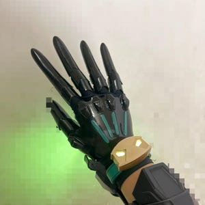 May include: A black articulated robotic glove featuring long, pointed finger segments, teal accents on the back of the hand, and a gold-coloured wrist cuff with two illuminated yellow lights. The design includes segmented plating.