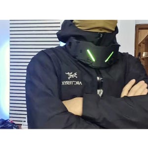 Pode incluir: Um protetor de pescoço tático preto com duas faixas de luz diagonais verde-brilhante na frente. O protetor é usado sobre uma jaqueta preta com capuz com o logotipo e texto Arc'teryx em branco no peito e um cachecol bege por baixo.
