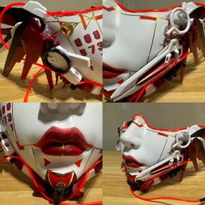 Może przedstawiać: Biała i czerwona maska ​​na twarz z misternymi detalami, w tym czerwono-białym skrzydłem, złotym zębatym kołem i czerwono-białym ostrzem. Maska ​​ma czerwone usta i białą twarz z czarnymi cyframi na czole. Maska ​​leży na drewnianej powierzchni.