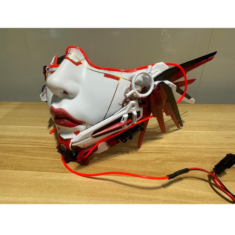 Może przedstawiać: Biała i czerwona maska ​​na twarz ze skomplikowanymi detalami i czerwonym kablem świetlnym. Maska ​​ma geometryczny wz&oacute;r z złotymi akcentami i czerwonym kablem świetlnym.