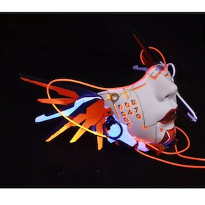 Może przedstawiać: Biała maska z pomarańczowymi i czarnymi akcentami i świecącymi pomarańczowymi i niebieskimi światłami. Maska ma geometryczny wz&oacute;r z liczbami i zębatką z boku.