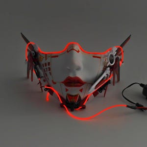 Może przedstawiać: Biała i czerwona maska ​​na twarz o futurystycznym designie. Maska ​​ma czerwone diody LED wok&oacute;ł krawędzi. Maska ​​jest wykonana z tworzywa sztucznego i ma szczeg&oacute;łowy projekt.