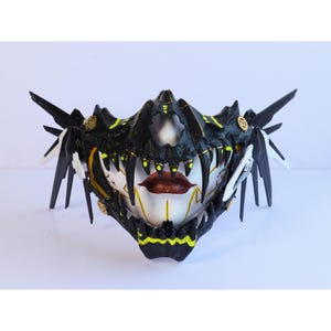 Op de afbeelding: Een zwart-wit masker met gele accenten. Het masker heeft een schedelvormig ontwerp met scherpe tanden en een gestileerd menselijk gezicht met rode lippen. Het masker heeft ingewikkelde details en een dramatische, bijna dreigende uitstraling.