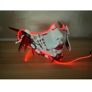 Może przedstawiać: Biała i czerwona maska ​​na twarz o futurystycznym designie. Maska ​​ma czerwone światło, kt&oacute;re oświetla twarz. Maska ​​ma wz&oacute;r liczbowy na czole.