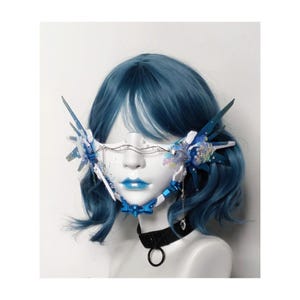 Puede incluir: Una máscara facial blanca con detalles azules y plateados. La máscara tiene un diseño geométrico y está decorada con lentejuelas iridiscentes. La máscara la lleva puesta una persona con el pelo azul.