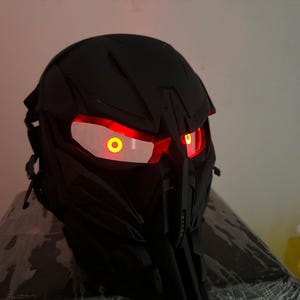 Puede incluir: Un casco negro con un diseño futurista. El casco tiene dos ojos rojos brillantes con círculos amarillos en el centro.