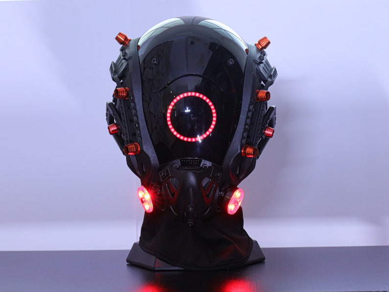 May include: A black helmet with a red ring light and red lights on the sides. The helmet is on a black stand.