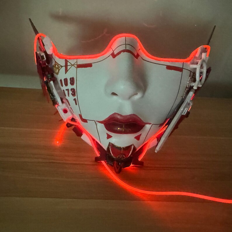 Może przedstawiać: Biała maska z czerwonymi akcentami i świecącym czerwonym światłem wok&oacute;ł krawędzi. Maska ma ludzką twarz z nosem, ustami i oczami. Maska jest wykonana z plastiku i ma futurystyczny design.