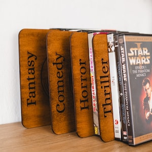 Puede incluir: Un conjunto de sujetalibros de madera marrón con las palabras "Fantasy", "Comedy", "Horror" y "Thriller". Junto a ellos, se encuentran carátulas de DVD, incluyendo "Star Wars", sobre una estantería de madera.