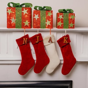 May include: Christmas decorations featuring three red gift box-shaped lights with green glitter accents and star cutouts. Below, four Christmas stockings hang, three red and one white, each with a wooden letter.