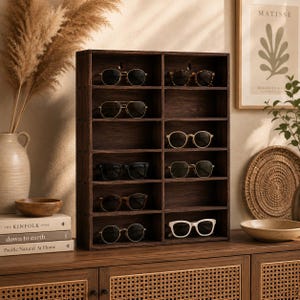 Puede incluir: Un estante de pared de madera de color marrón oscuro con doce compartimentos rectangulares individuales. El organizador contiene varios estilos de gafas y gafas de sol, incluyendo monturas negras, blancas y de carey dispuestas en dos columnas verticales.