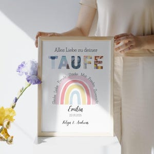 Personalisierbares Geldgeschenk Taufe Mädchen mit Geld in einem hellen Holzbilderrahmen steht auf einem weißen Tisch und wird von einer Frau in heller Kleidung gehalten. An dem Bilderrahmen lehnt eine gelb-lila Blume.
