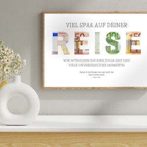 Geldgeschenk Reisekasse in einem Holzbilderrahmen hängt an einer beigen Wand über einem Sideboard. Auf dem Sideboard stehen eine Pflanze in einem gelben Blumentopf sowie eine weiße Vase.