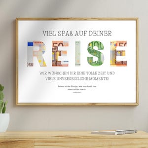Geldgeschenk Reisekasse in einem Holzbilderrahmen hängt an einer beigen Wand über einem Sideboard. Daneben hängt eine Lampe und auf dem Sideboard steht ein Kaktus und liegt ein Notizbuch. Neben dem Sideboard steht eine weitere Pflanze.