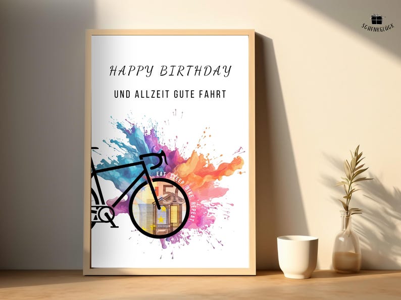 Geldgeschenk Fahrrad Geburtstag in einem Holzbilderrahmen auf dem Boden vor einer beigen Wand neben einer weißen Vase und einer Glasvase mit einem grünen Zweig darin.