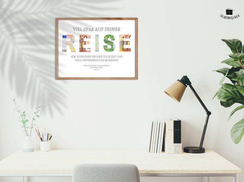 Geldgeschenk Reisekasse in einem Holzbilderrahmen hängt an einer weißen Wand über einem Schreibtisch. Auf dem Schreibtisch stehen eine Pflanze, Stif, Bücher sowie eine Lampe. Daneben steht eine Pflanze und davor ein Schreibtischstuhl.