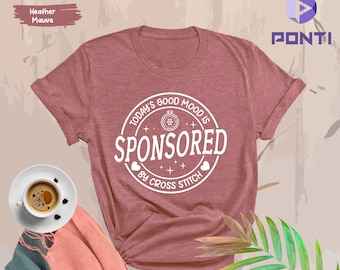 May include: A heather mauve t-shirt with a white circle graphic that says "Today's Good Mood Is Sponsored By Cross Stitch".