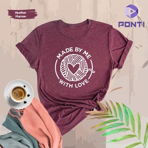 May include: A heather maroon t-shirt with a white graphic of a ball of yarn with a heart in the centre and the text "Made by me with love".