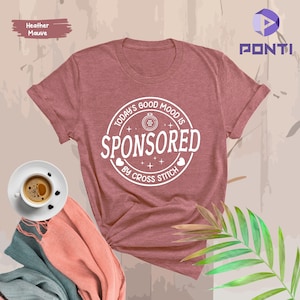 May include: A heather mauve t-shirt with a white circle graphic that says "Today's Good Mood Is Sponsored By Cross Stitch".