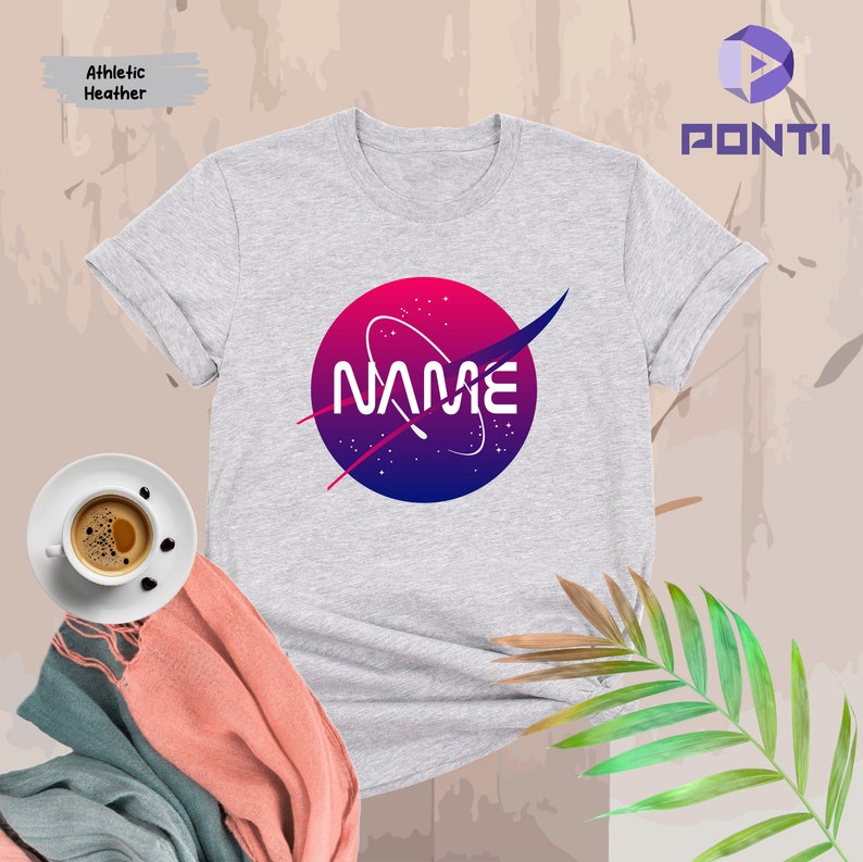Może przedstawiać: Szary T-shirt z logo inspirowanym NASA w kolorze r&oacute;żowym, niebieskim i białym, na kt&oacute;rym widnieje napis "NAME".