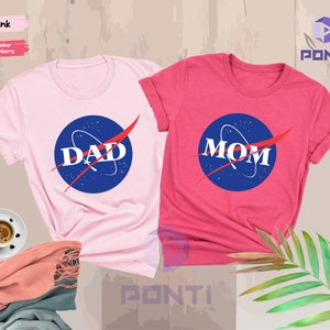 K&ouml;nnte beinhalten: Zwei T-Shirts, eines rosa und eines rot, mit einem NASA-inspirierten Logo, auf dem "DAD" und "MOM" in roten Buchstaben stehen.