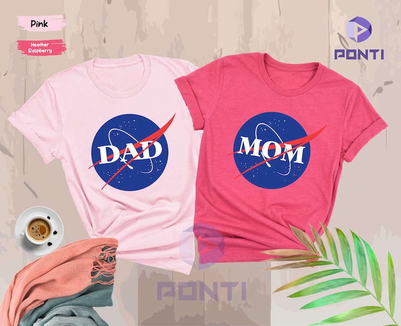 K&ouml;nnte beinhalten: Zwei T-Shirts, eines rosa und eines rot, mit einem NASA-inspirierten Logo, auf dem "DAD" und "MOM" in roten Buchstaben stehen.
