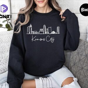 May include: A black sweatshirt with a white outline of the Kansas City skyline and the text "Kansas City" in white cursive.