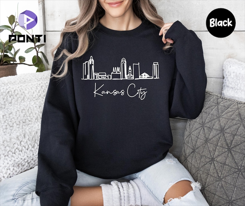 May include: A black sweatshirt with a white outline of the Kansas City skyline and the text "Kansas City" in white cursive.