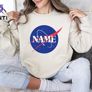 K&ouml;nnte beinhalten: Ein sandfarbener Pullover mit einem blauen und roten NASA-inspirierten Logo, auf dem "NAME" in roten Buchstaben steht.