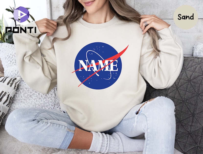 K&ouml;nnte beinhalten: Ein sandfarbener Pullover mit einem blauen und roten NASA-inspirierten Logo, auf dem "NAME" in roten Buchstaben steht.