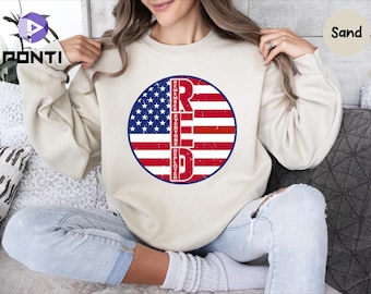 May include: A white sweatshirt with a red, white, and blue American flag design. The flag is in a circle with the word "RED" in large letters and the words "REMEMBER EVERYONE DEPLOYED" in smaller letters around the flag.