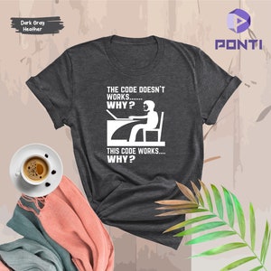 May include: Dark gray heather t-shirt with white text and graphic. The text reads "THE CODE DOESN'T WORKS... WHY? THIS CODE WORKS... WHY?" A graphic depicts a person at a computer. The shirt is a casual wear item.