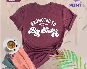 May include: A heather maroon t-shirt with the text "Promoted to Big Sister" in white with a heart and arrow design.
