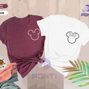 以下が含まれることがあります： 2つの半袖Tシャツ。1つはヘザーマルーン、もう1つは白。各シャツにはミッキーマウスの頭の輪郭があり、1つはリボン付き。シャツは、コーヒーカップとスカーフのあるニュートラルな背景に表示されています。
