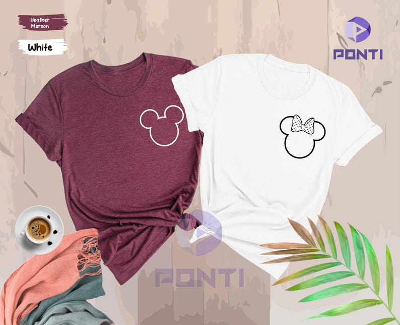以下が含まれることがあります： 2つの半袖Tシャツ。1つはヘザーマルーン、もう1つは白。各シャツにはミッキーマウスの頭の輪郭があり、1つはリボン付き。シャツは、コーヒーカップとスカーフのあるニュートラルな背景に表示されています。