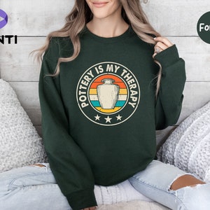 May include: A dark green sweatshirt with a vintage-style graphic that says "Pottery is my therapy" with a stylized image of a pottery vase.
