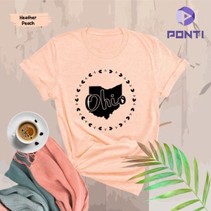 K&ouml;nnte beinhalten: Ein T-Shirt in der Farbe Heidepfirsich mit einer schwarzen Grafik des Bundesstaates Ohio, auf dem das Wort "Ohio" in einer stilisierten Schriftart innerhalb der Staatsgrenze steht. Die Grafik ist von einem Kreis aus kleinen Herzen umgeben.