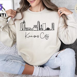 May include: A sand-colored sweatshirt with a black outline of the Kansas City skyline and the text "Kansas City" in cursive.