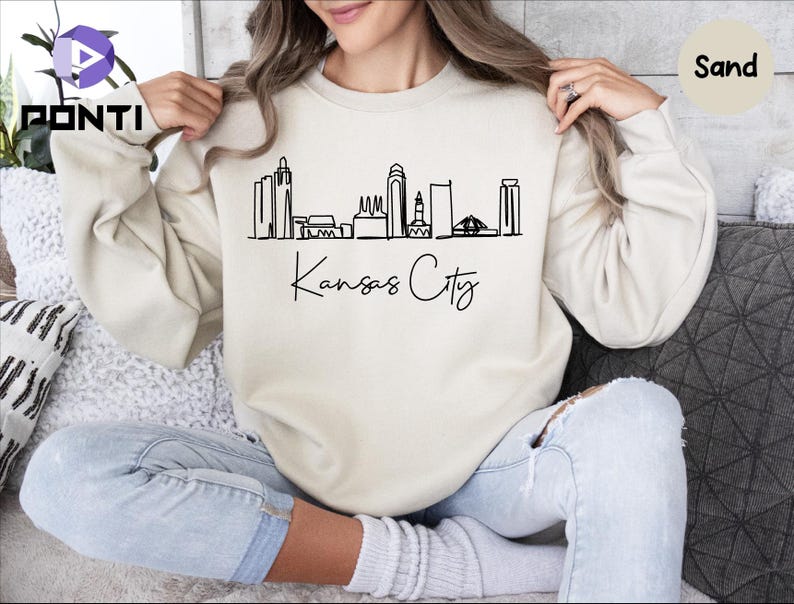 May include: A sand-colored sweatshirt with a black outline of the Kansas City skyline and the text "Kansas City" in cursive.