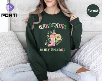 May include: A dark green sweatshirt with the text "GARDENING is my therapy" in rainbow colors. A pink gardening boot with flowers is pictured next to the text.