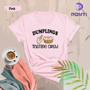 Może przedstawiać: R&oacute;żowa koszulka z napisem "Dumplings Tasting Crew" i kresk&oacute;wkowym obrazkiem pierog&oacute;w w koszyku.