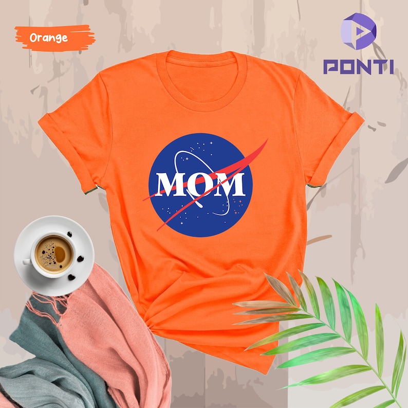 K&ouml;nnte beinhalten: Orangefarbenes T-Shirt mit einem blauen und roten NASA-inspirierten Logo, auf dem "MOM" in wei&szlig;en Buchstaben steht.