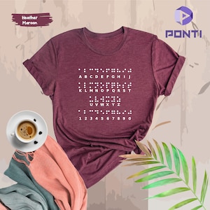 May include: A heather maroon t-shirt with a white print of the alphabet and numbers in braille. The text reads: 'ABCDEFGHIJ KLMNOPQRST UVWXYZ 1 2 3 4 5 6 7 8 9 0'.