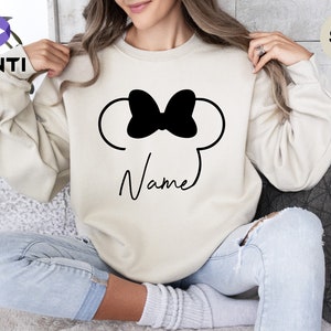 May include: Sand-colored sweatshirt featuring a black bow and mouse ears design. The word "Name" is written in a cursive font below the design. The sweatshirt is a neutral color and has long sleeves.