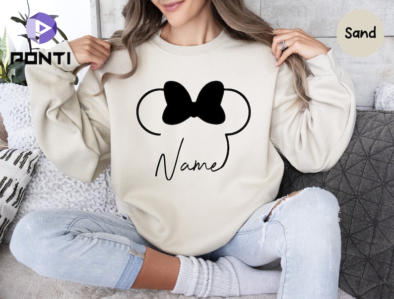 May include: Sand-colored sweatshirt featuring a black bow and mouse ears design. The word "Name" is written in a cursive font below the design. The sweatshirt is a neutral color and has long sleeves.
